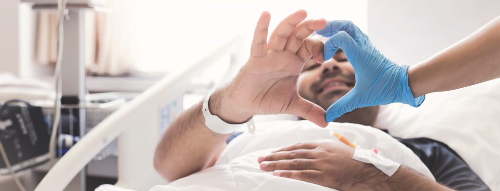 Un soignant et un patient dans un lit d’hôpital joignent leurs mains pour former un cœur avec leurs doigts.
