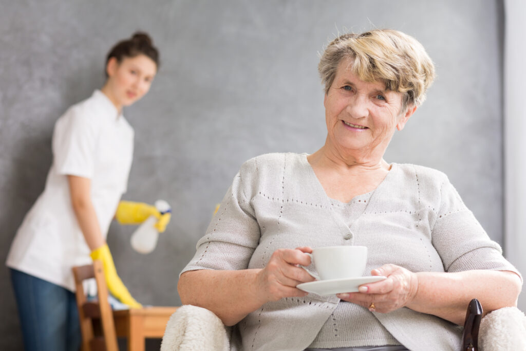Une dame âgée souriante sirote son thé pendant qu’un aide soignant l’aide à nettoyer sa maison